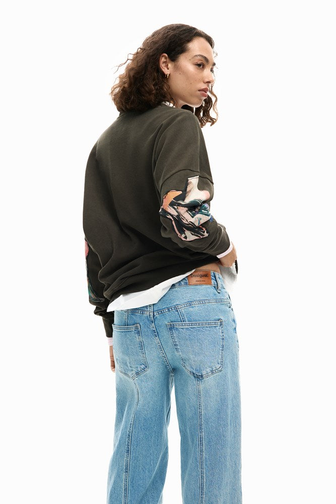 Person wearing a dark green sweatshirt with colorful designs and light blue jeans on a white background