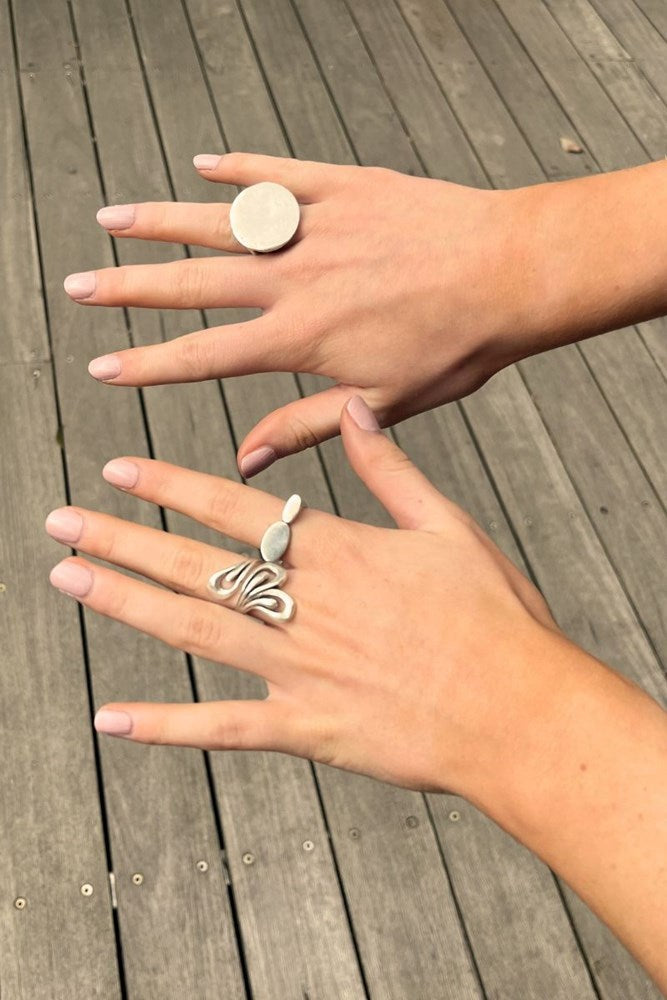 Two hands displaying silver rings on a weathered gray wooden deck. The top hand wears a large, round disc-shaped silver ring, and the bottom hand wears a silver ring with a wavy, abstract design and a small light blue stone.