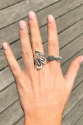 A silver ring with an abstract, wavy openwork design on one finger and a small flat oval stone on another finger, shown on a hand against a gray wooden deck background.