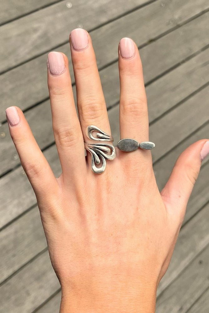 A silver ring with an abstract, wavy openwork design on one finger and a small flat oval stone on another finger, shown on a hand against a gray wooden deck background.