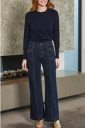 Woman wearing a navy sweater and dark blue jeans standing in a room with a stone wall.