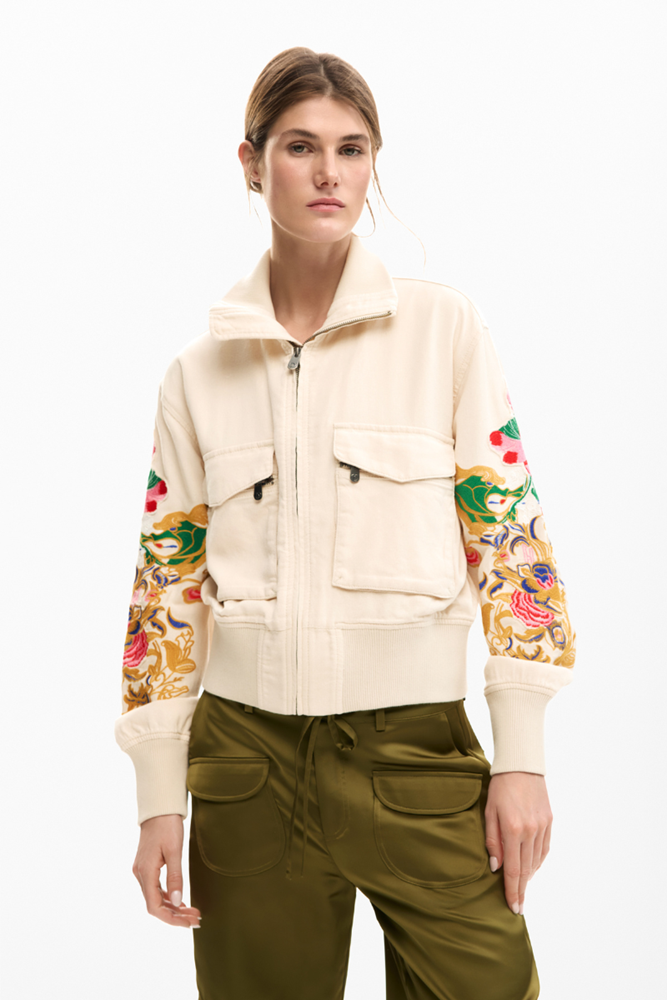 A model wearing a cream-colored zip-up jacket with colorful embroidered sleeves and olive green pants, shot against a plain white background.