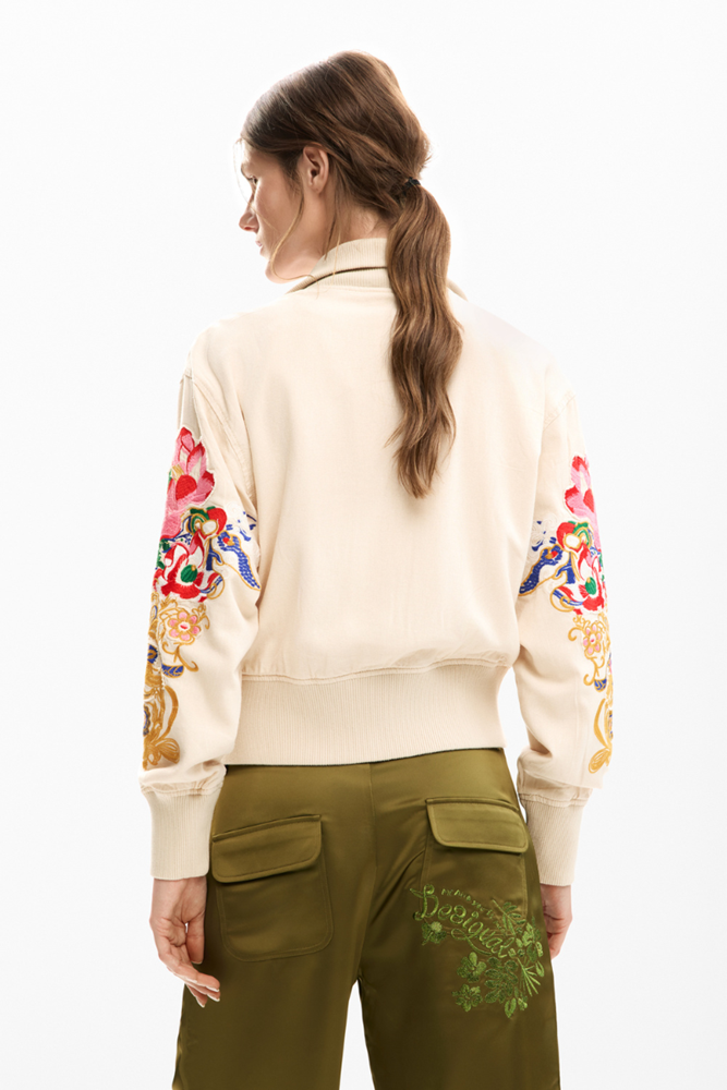 Person wearing a cream jacket with floral designs on sleeves and green pants with floral patterns, on a white background.