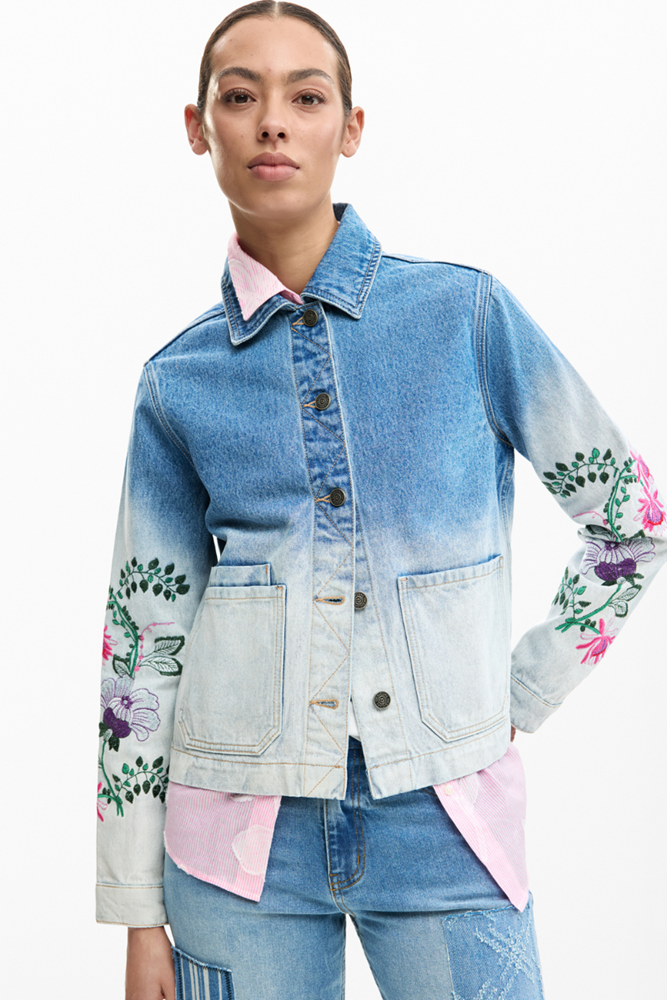 Person wearing a denim jacket with floral embroidery on a white background