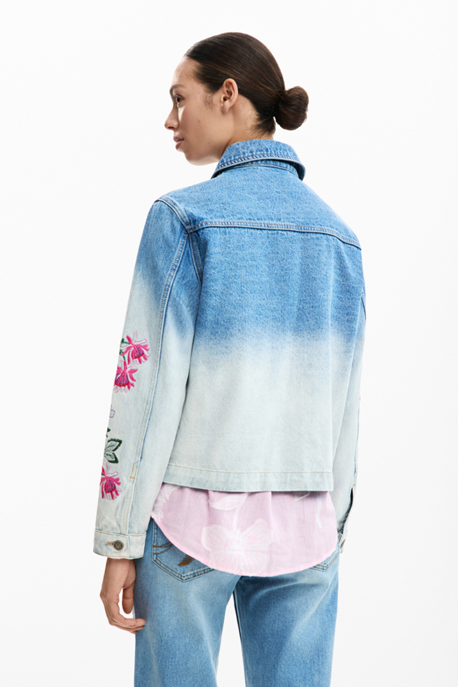 Woman wearing a blue gradient denim jacket with floral embroidery on a white background