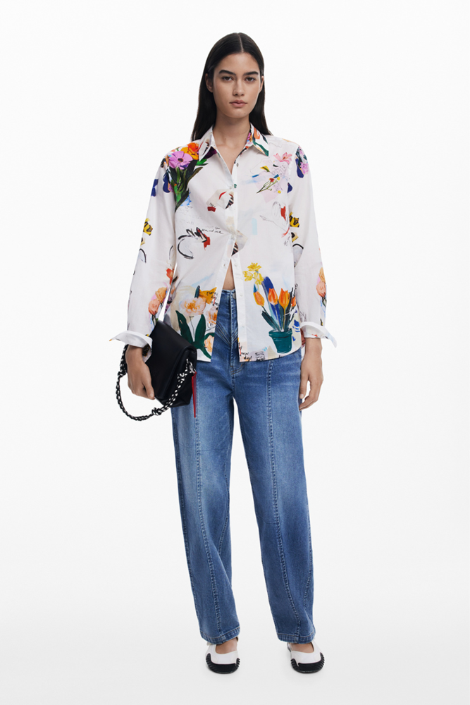 A model stands against a plain white studio background wearing a white long-sleeve button-up shirt with a colorful floral and bird print, blue jeans, black-and-white shoes, and holding a small black handbag.