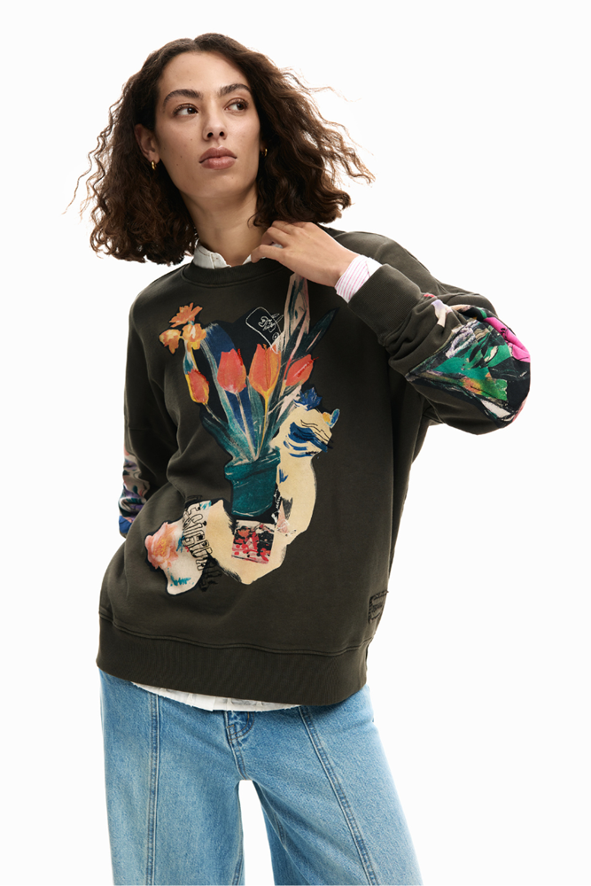 A woman wearing a dark olive green crewneck sweatshirt with a colorful graphic print of a hand holding a bouquet of flowers, paired with light blue jeans, against a plain white background.