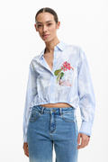 Woman wearing a light blue shirt with floral embroidery and high-waisted jeans on a white background