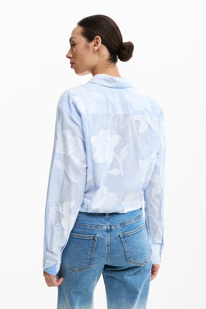 A woman shown from the back wearing a light blue long-sleeve button-up shirt with a white floral pattern and blue jeans, against a plain white studio background.