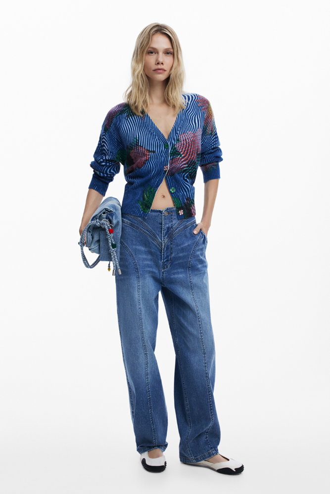 A model stands against a plain white studio background wearing a blue patterned cropped cardigan, wide-leg blue jeans, and black-and-white flats, while holding a small blue denim bag.