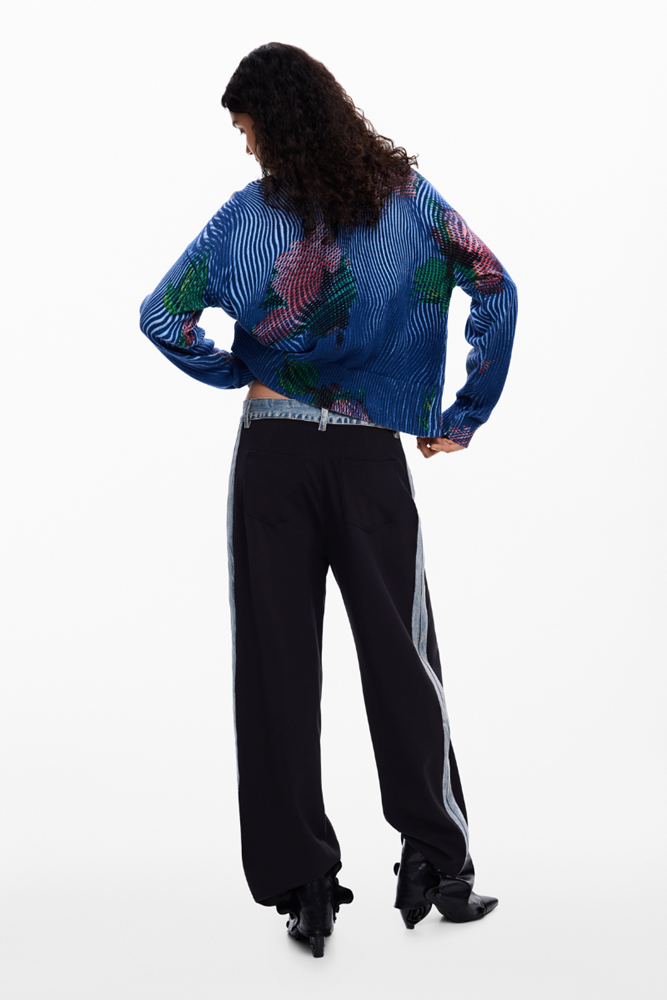 A model is shown from the back wearing a blue patterned long-sleeve crop top and black wide-leg track pants with white side stripes, set against a plain white studio background.