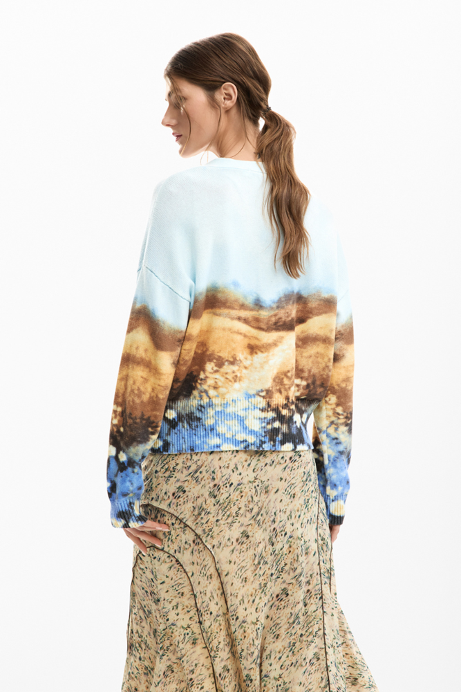 A woman shown from the back wearing a long-sleeve crewneck sweatshirt with a scenic landscape print in blue, brown, and beige, paired with a beige floral print skirt, against a plain white background.