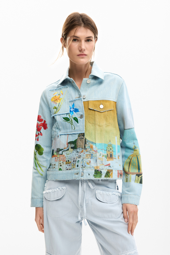 A model wearing a light blue denim jacket with colorful, artistic prints on the front and sleeves, paired with matching light blue denim pants, against a plain white background.