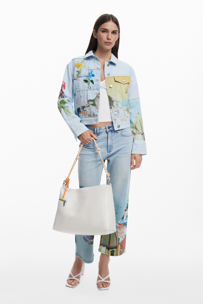 A model stands against a white studio background wearing a light blue cropped denim jacket with colorful floral and scenic prints, matching light blue jeans with multicolor printed cuffs, a white crop top, white strappy heels, and holding a large off-white tote bag.