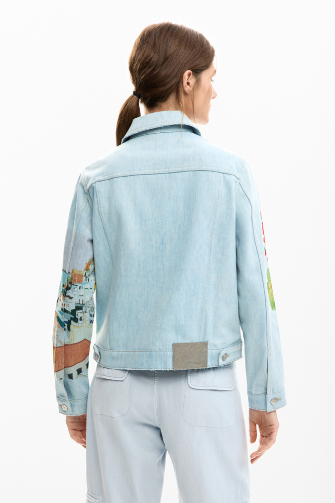 A model shown from the back wearing a light blue denim jacket with colorful patchwork illustrations on the sleeves and matching light blue jeans against a plain white background.