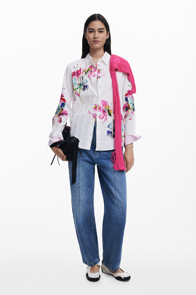 A model stands against a plain white studio background wearing a white long-sleeve button-up shirt with colorful floral prints, blue jeans, and black-and-white flats. A bright pink scarf is draped over her shoulder, and she holds a small black clutch.