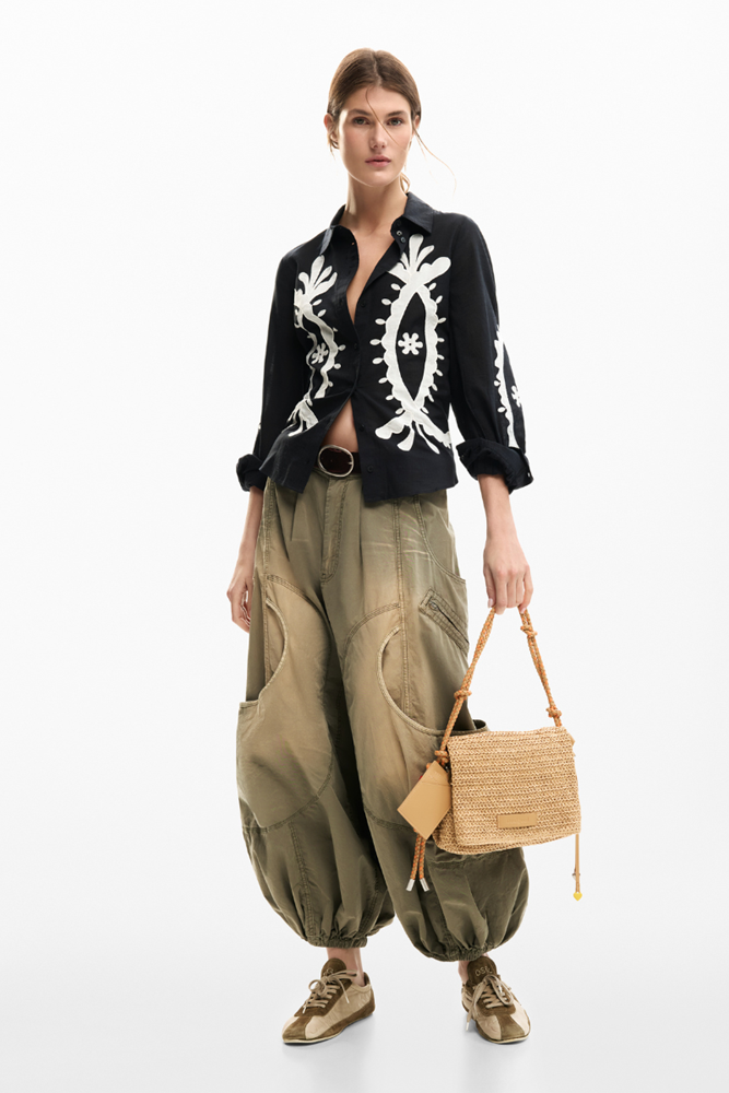 A model stands against a white studio background wearing a black cropped button-up shirt with white embroidered patterns, loose olive green baggy pants, and carrying a small woven straw handbag.