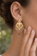 Gold earrings with a central gemstone worn by a person, with a blurred natural background.