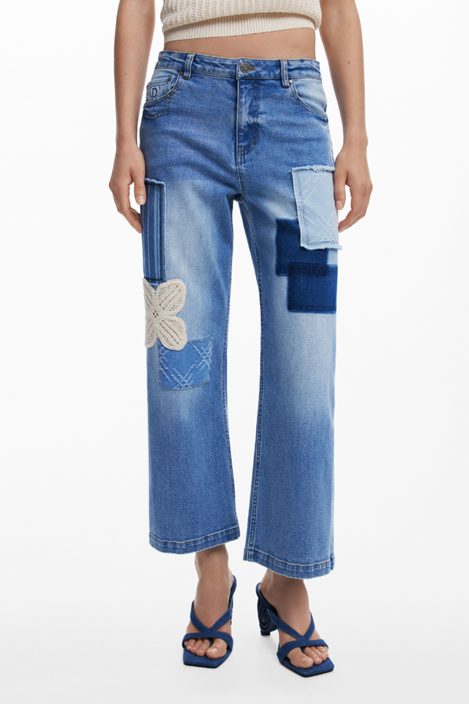A model wearing a pair of cropped wide-leg blue denim jeans with patchwork details, including a large light blue square patch on the thigh and a white embroidered floral motif, paired with blue strappy heels.