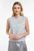 A blonde woman wearing a sleeveless silver sequined top with gray ribbed trim and a matching gray metallic skirt, photographed against a plain light gray studio background.
