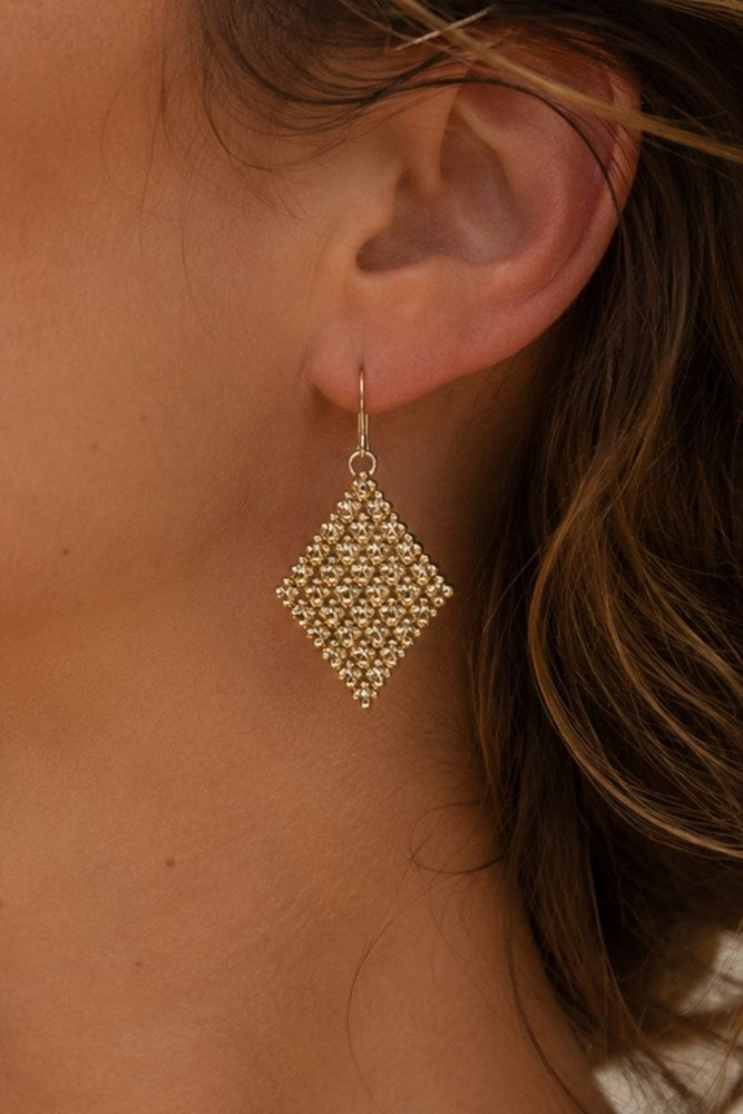 A close-up of a person's ear wearing a dangling geometric beaded earring in a warm metallic gold tone.
