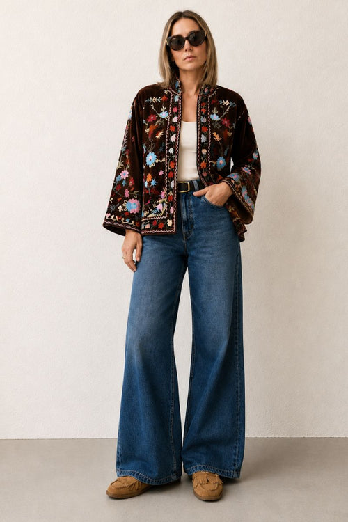Woman wearing a patterned jacket, white top, blue jeans, and brown shoes against a plain background
