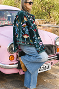 Person wearing a floral embroidered jacket and jeans standing next to a pink vintage car.