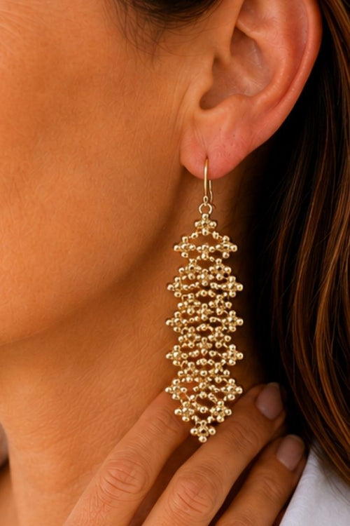 A close-up of a model wearing a long, dangling gold beaded earring. The earring features a cluster of small gold beads forming a textured, tiered design hanging from a gold fishhook earwire.