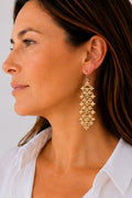 A close-up side profile of a woman wearing a long, cascading gold beaded dangle earring against a plain light background.