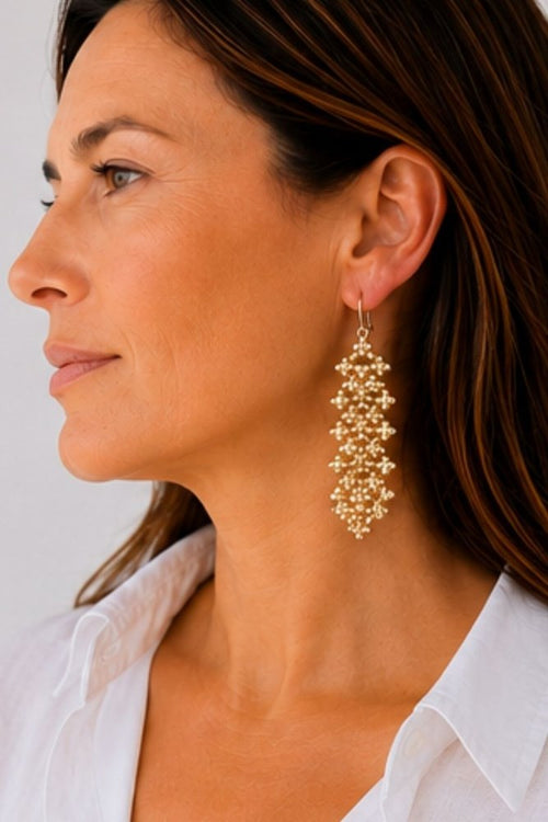 A close-up side profile of a woman wearing a long, cascading gold beaded dangle earring against a plain light background.