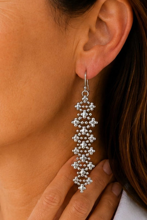 A close-up of a woman's ear wearing a long, dangling silver earring with a textured, beaded geometric design.