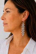 A close-up side profile of a woman wearing a long, dangling silver-toned earring with multiple small clear stones arranged in a floral or snowflake pattern.