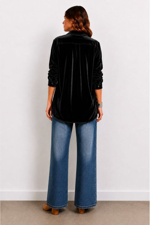 Person wearing a black velvet shirt and blue jeans against a white wall.