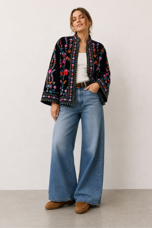Woman wearing a patterned jacket, white top, blue jeans, and brown shoes on a plain background