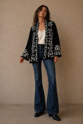 Woman wearing a black embroidered jacket, white top, and blue jeans against a beige background