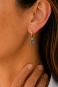 A close-up of a model's ear wearing a small gold huggie hoop earring with a rectangular blue gemstone.