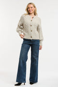 A woman modeling a light beige cable-knit cardigan sweater with brown buttons, paired with dark blue wide-leg jeans and black heels, against a plain white studio background.