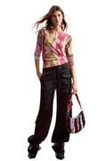 A model wearing a pink and gold patterned long-sleeve top, black wide-leg cargo pants, and black boots, holding a small patterned handbag against a white background.