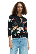 A woman wearing a black crewneck long-sleeve sweater with a vibrant multicolor floral and abstract print, styled with light wash blue jeans and a black belt, against a plain white background.