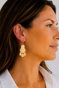 A close-up side profile of a woman wearing a pair of gold dangling earrings with a layered, flower-like design, paired with a white collared shirt against a light gray background.