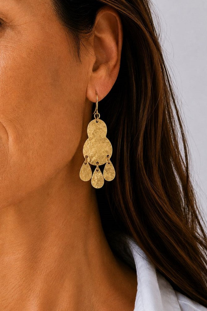 A close-up of a woman's ear wearing a textured gold dangling earring with three teardrop-shaped charms, against a light gray background.