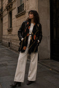 Woman wearing a black leather jacket with orange patterns, white pants, and black shoes on a city street.