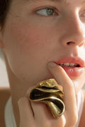 A close-up lifestyle shot of a model wearing a large, organic-shaped gold ring on her finger, with her hand resting near her mouth.