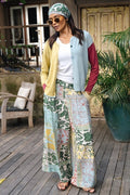 A woman stands on a wooden deck wearing a colorful patchwork wide-leg pants set with a white top, a light blue and red cardigan, a green patterned headscarf, and sunglasses.