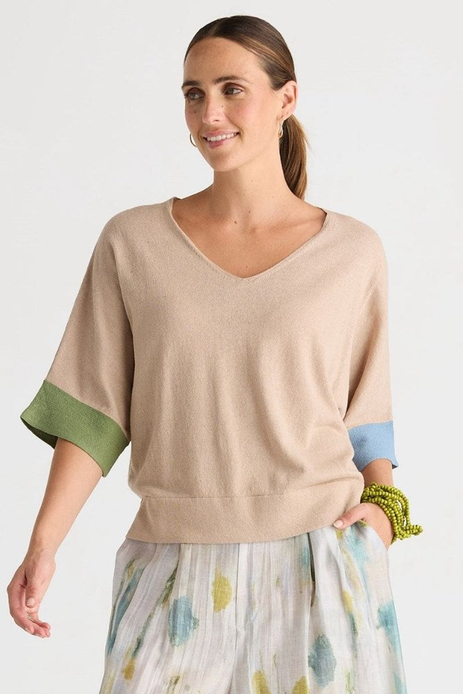 A woman wearing a beige knit sweater with green and light blue color-blocked elbow-length sleeves, paired with a white skirt featuring a blue and yellow floral pattern, against a plain light gray background.