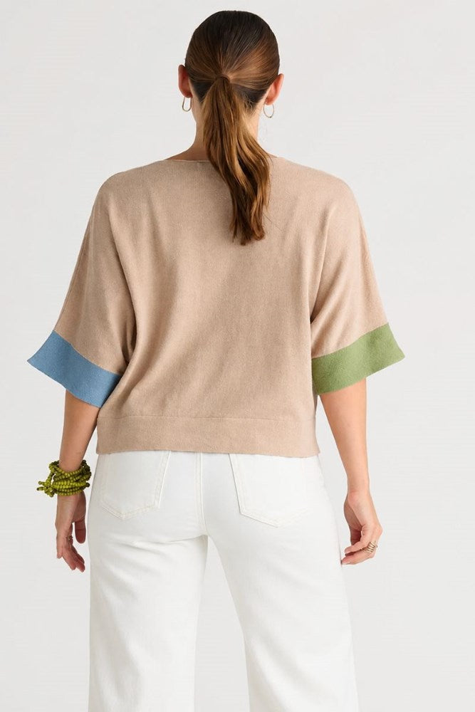 A woman shown from the back wearing a beige knit sweater with contrasting light blue and sage green sleeve cuffs, paired with white pants, against a plain light gray studio background.