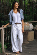 A woman stands on a wooden deck in a tropical outdoor setting, wearing a white sleeveless top and white wide-leg pants with a brown belt, layered with a blue and white patterned short-sleeve cardigan.
