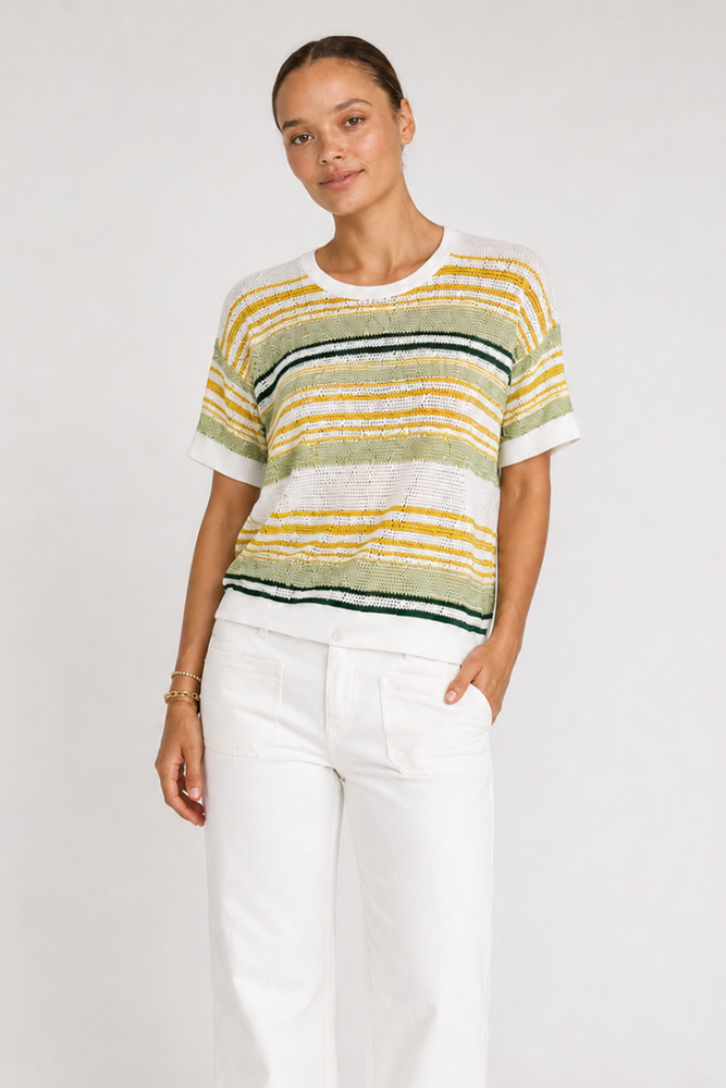 A woman modeling a short-sleeve knit top with horizontal stripes in yellow, sage green, white, and dark green, paired with white pants, against a plain light gray background.