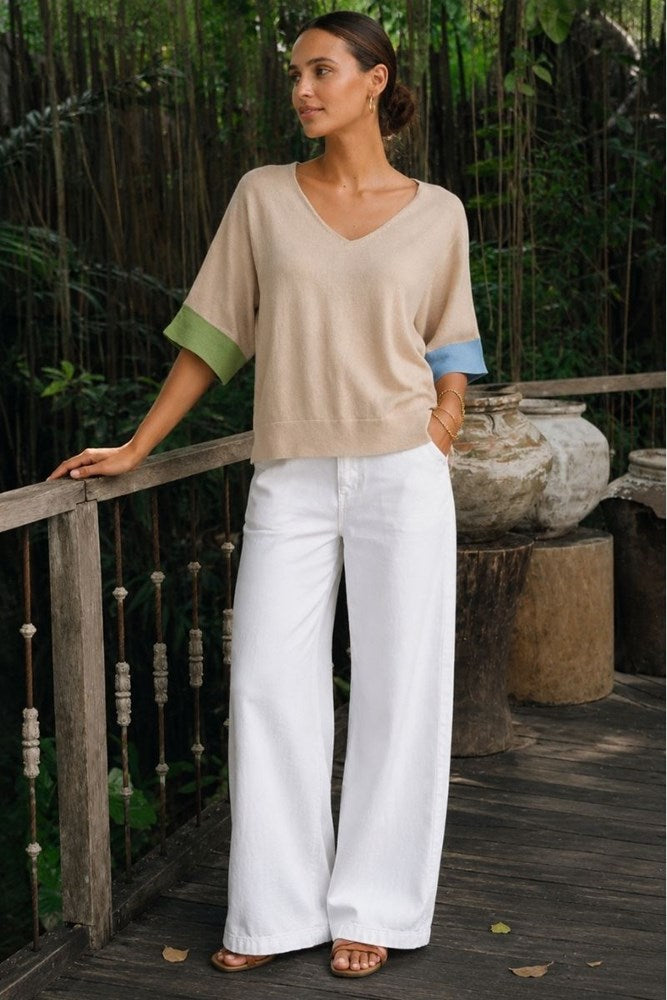 A woman stands on a wooden deck in a tropical garden setting, wearing a beige knit V-neck top with green and blue color-blocked elbow-length sleeves, paired with white wide-leg pants.