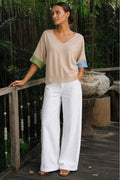 A woman stands on a wooden deck in a tropical garden setting, wearing a beige knit V-neck top with green and blue color-blocked elbow-length sleeves, paired with white wide-leg pants.